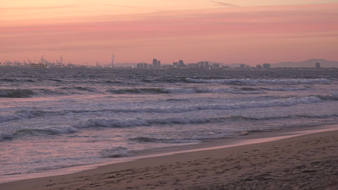 Huntington Beach waves spread across the midground as the wet sand catches pink light, with distant skyline shapes and no visible people.
