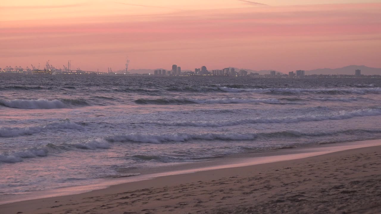 A fuller band of whitewater crosses the Huntington Beach shoreline beneath a pink and lavender sky, with cranes and buildings faint in the distance and no visible people.