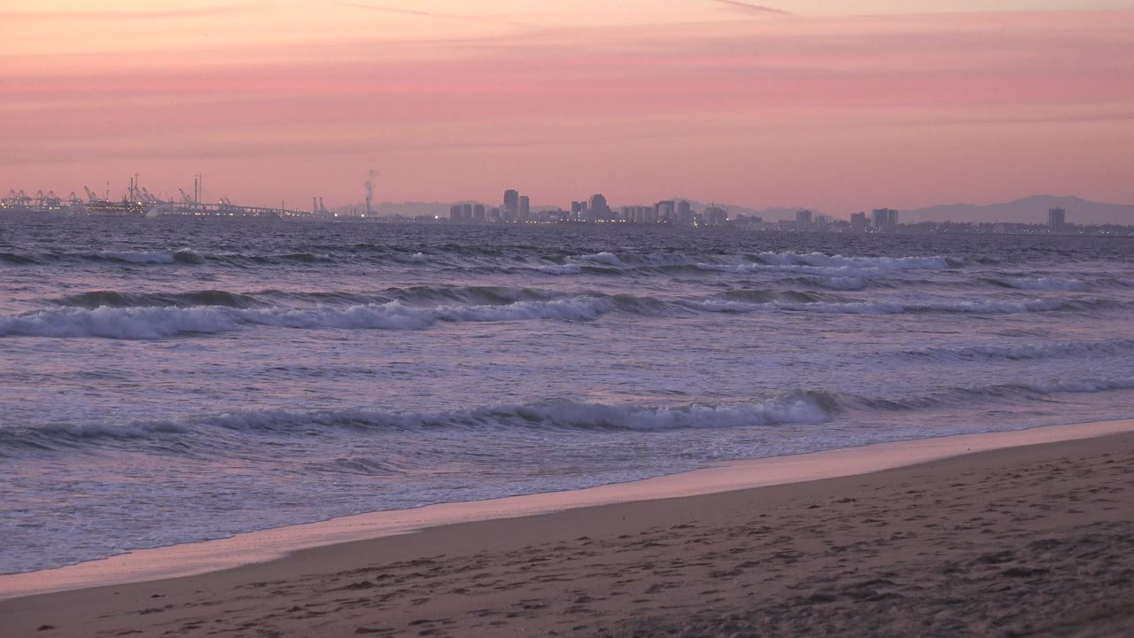 Huntington Beach shoreline with empty sand in the foreground, rolling waves offshore, a distant horizon skyline, and no visible people at the start of the clip.
