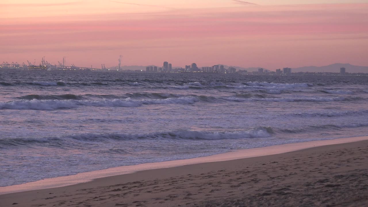 Huntington Beach surf rolls toward a clear strip of empty sand, with muted pink daylight over the water and no visible people in frame.