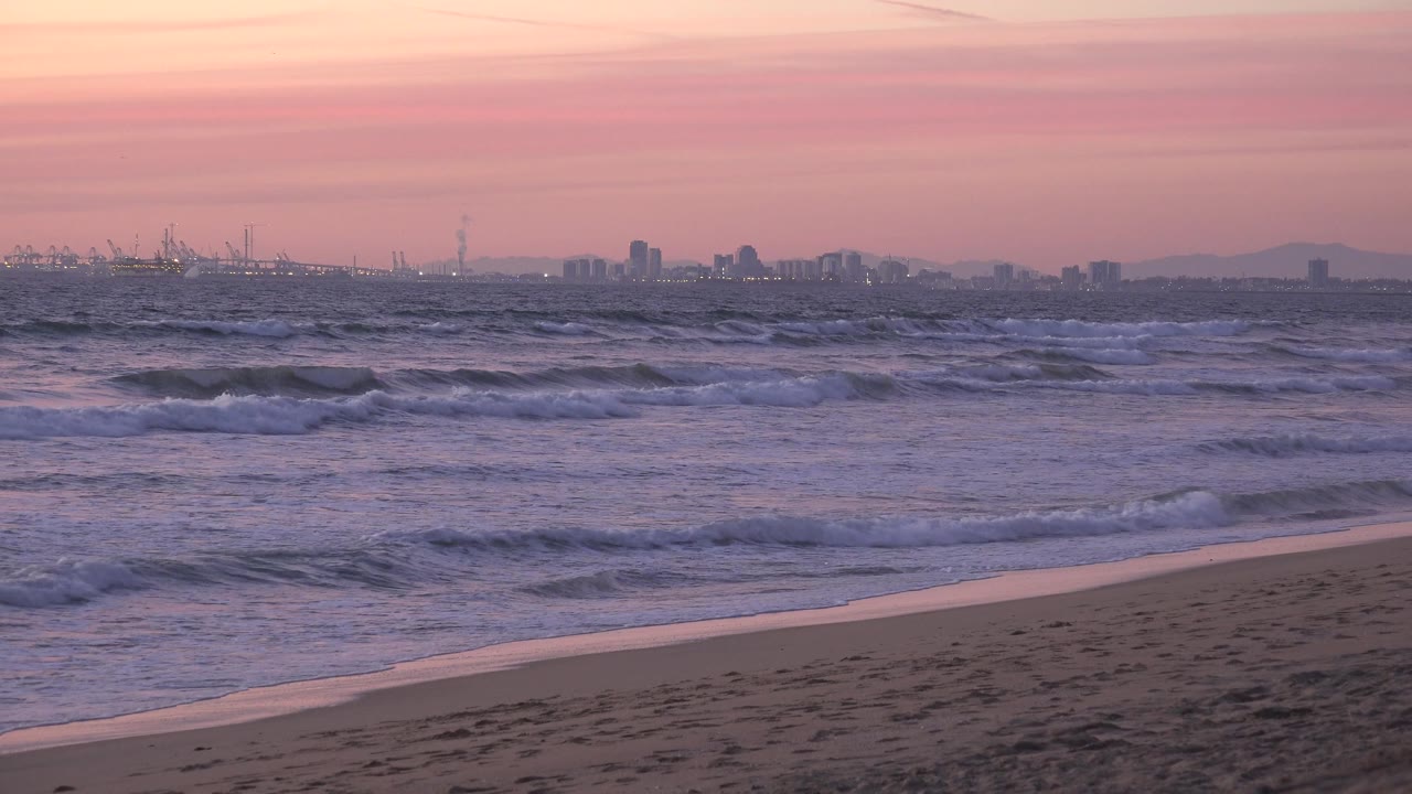 A sloped empty beach cuts across the lower frame at Huntington Beach, while broken waves fill the middle distance and a faint skyline sits on the horizon with no visible people.