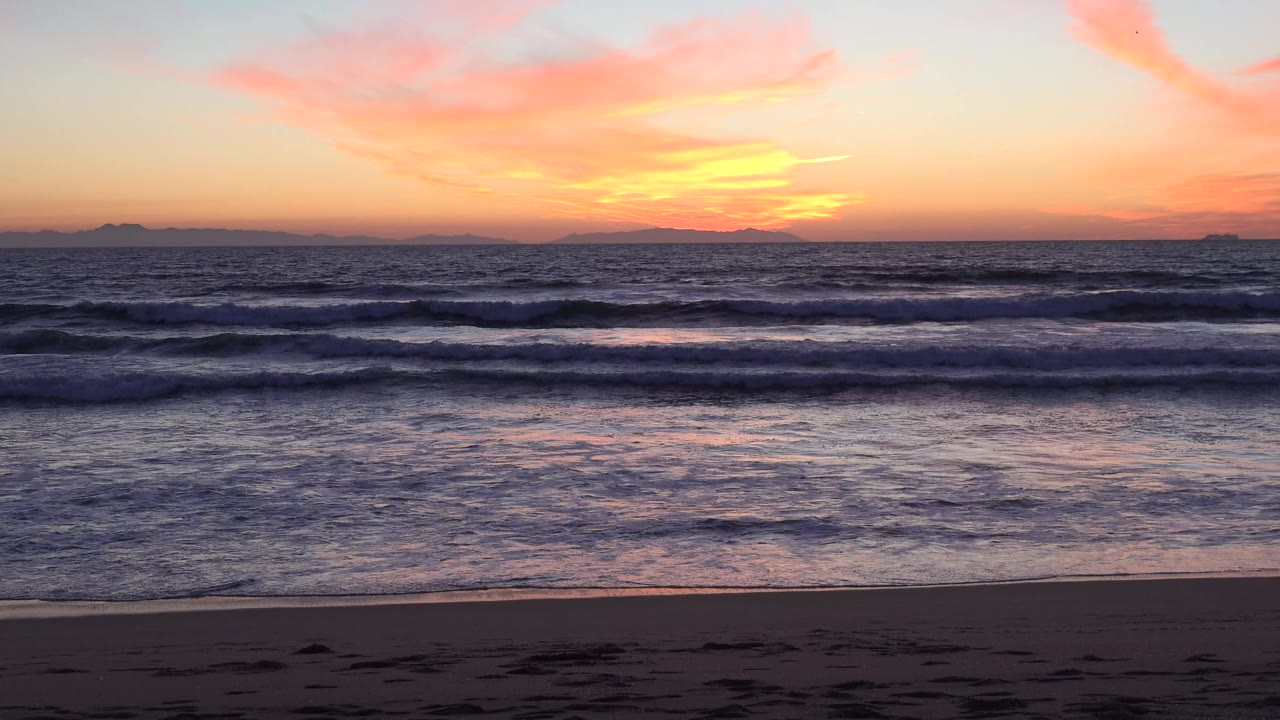 Huntington Beach sunset over low Pacific waves, dark sand in the foreground, distant horizon shapes, and no visible people.