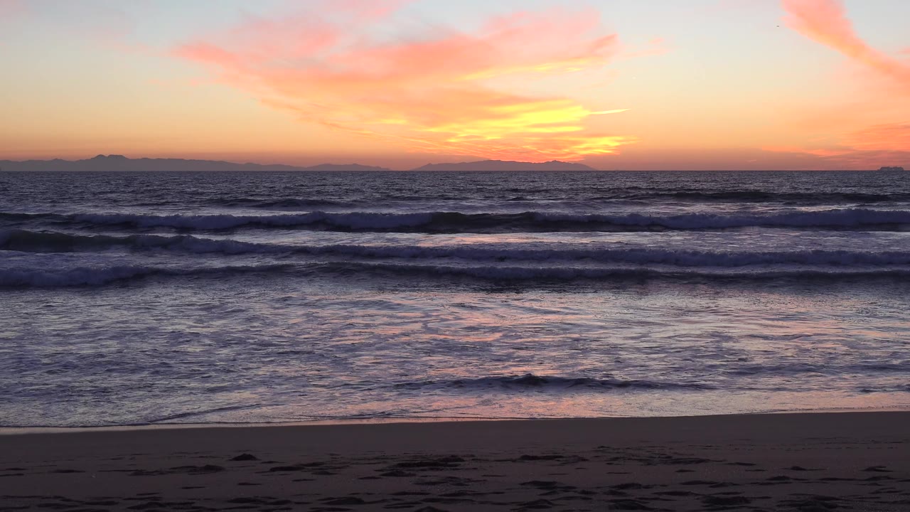 Huntington Beach shoreline at sunset with layered wave breaks across the midground, pink cloud streaks overhead, and no visible people.