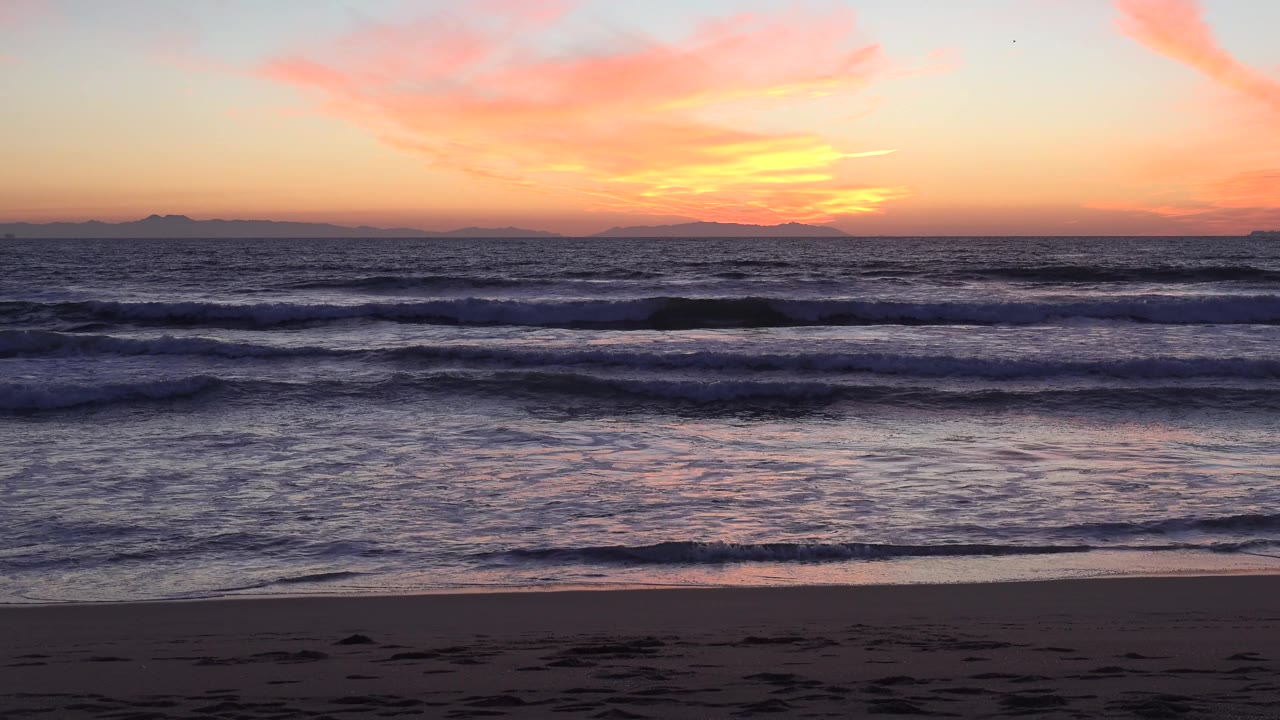Huntington Beach sunset scene with a darker foreground beach, white foam at the waterline, and no visible people in the frame.