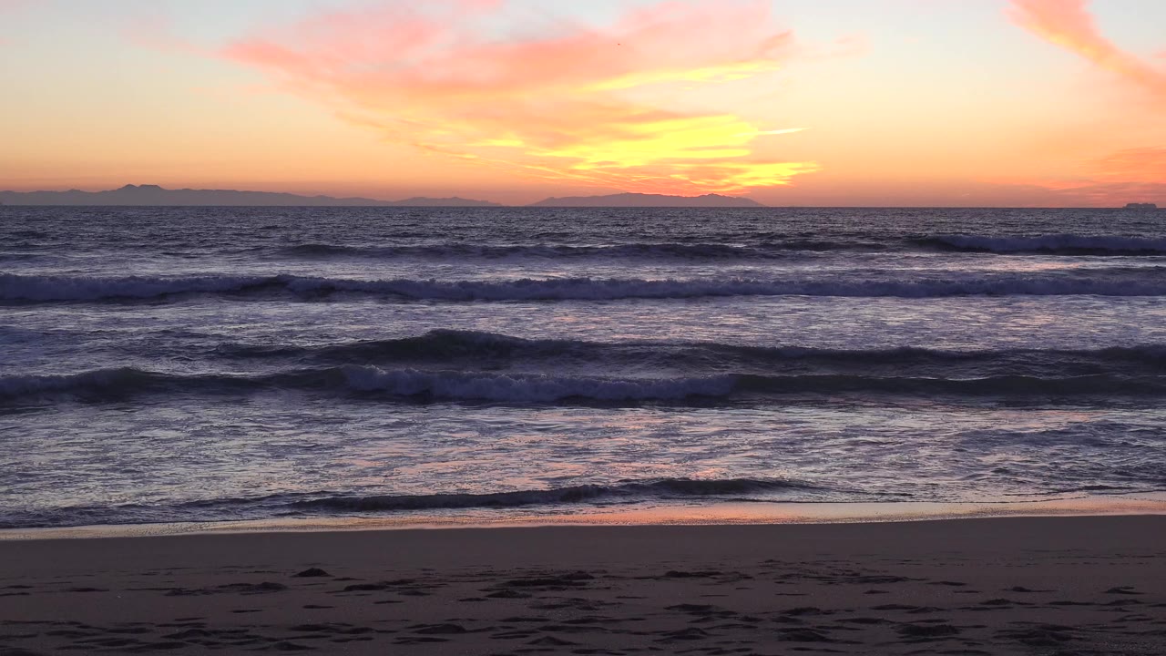 Opening frame of Huntington Beach at sunset with orange and pink sky above layered surf, dark sand in the foreground, distant land on the horizon, and no visible people.