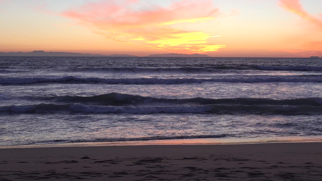 Huntington Beach ocean scene with darker blue waves breaking in parallel lines, warm sunset color reflected on the water, sandy foreground, and no visible people.
