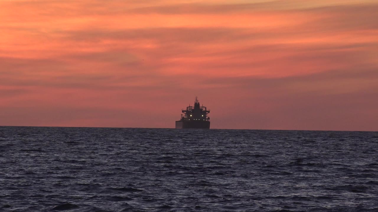 A distant transport boat sits near the center horizon off Huntington Beach, California, with dark ocean water in front, orange sunset clouds behind it, and no visible people.
