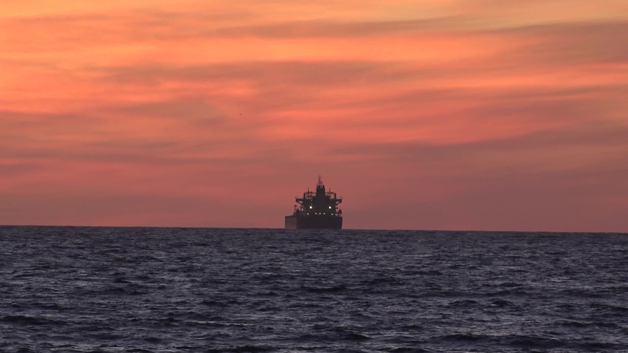 The transport boat appears farther right on the Huntington Beach ocean horizon, backed by red-orange cloud layers above choppy water and no visible people.