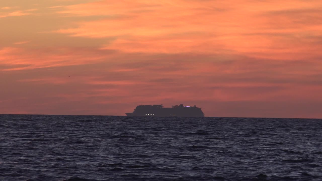 At 0:01 in Huntington Beach, the cruise ship remains near the center of the horizon beneath streaked orange cloud layers, with dark textured water in the foreground and no visible people.