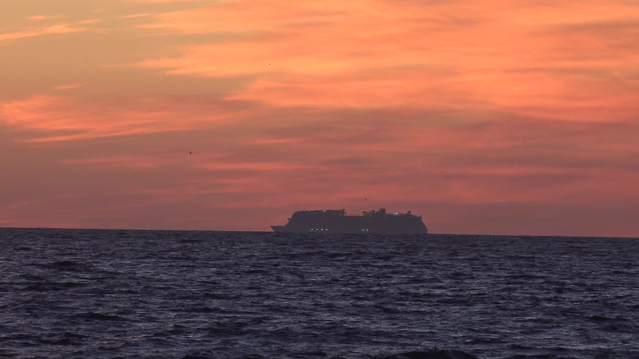 At 0:02 in Huntington Beach, the cruise ship appears farther right on the horizon line, silhouetted beneath a broad orange-pink sky with rippled dark ocean in front and no visible people.