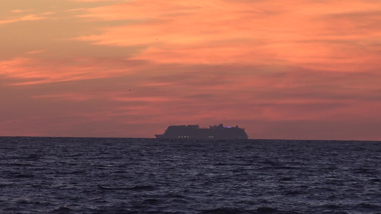 At 0:03 in Huntington Beach, the ship sits low and left of center against the horizon, with a wide band of dark ocean below and soft orange cloud texture overhead, and no visible people.