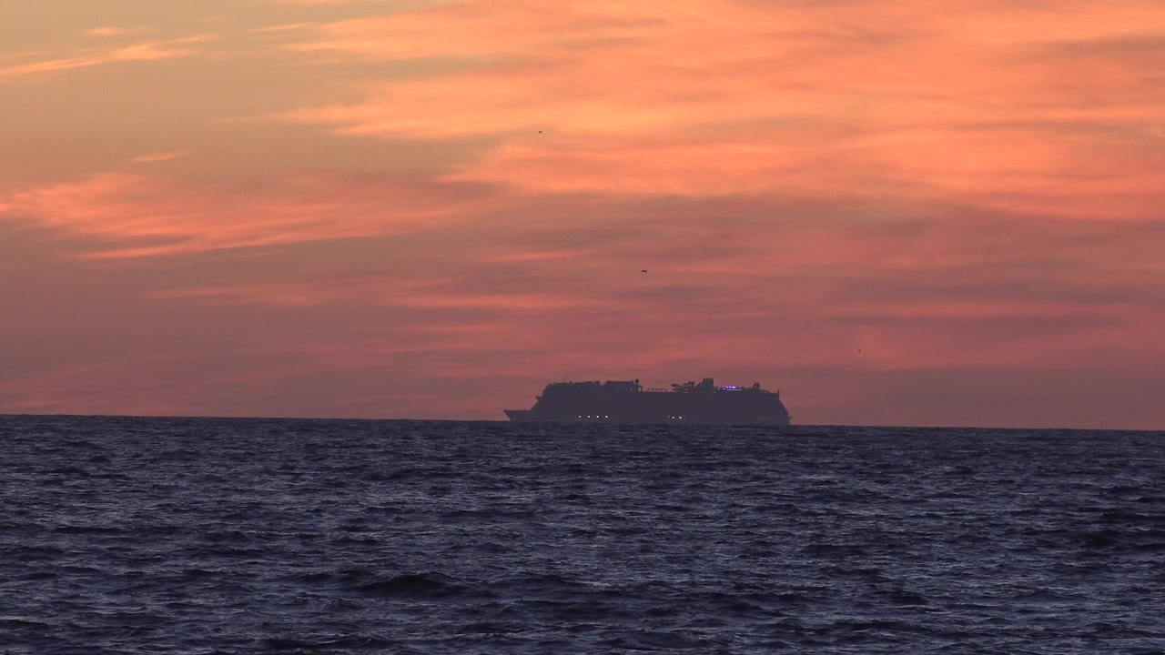 A distant cruise ship crosses the horizon off Huntington Beach, California, with dark rippled water below orange clouds and no visible people.