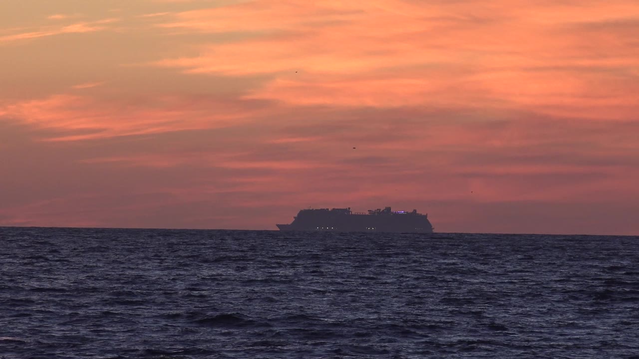 The cruise ship remains near the low horizon off Huntington Beach, California, with textured dark water in the foreground and no visible people.