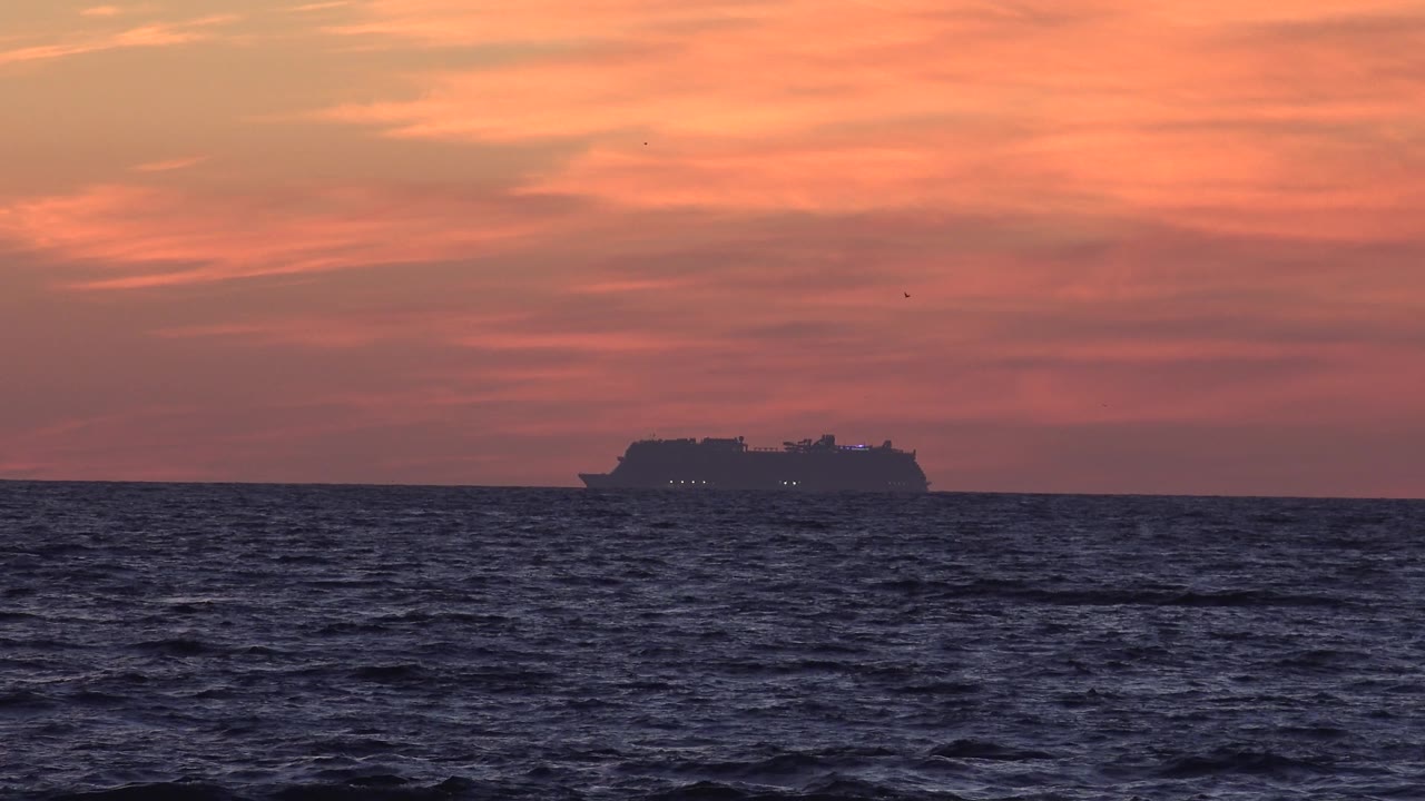 The vessel appears low and distant on the Huntington Beach horizon while small waves pattern the foreground and the sky stays orange, with no visible people.