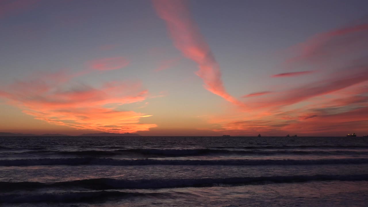 Huntington Beach, California sunset view with dark rolling waves in the foreground, pink clouds above the horizon, distant ship silhouettes, and no visible people.