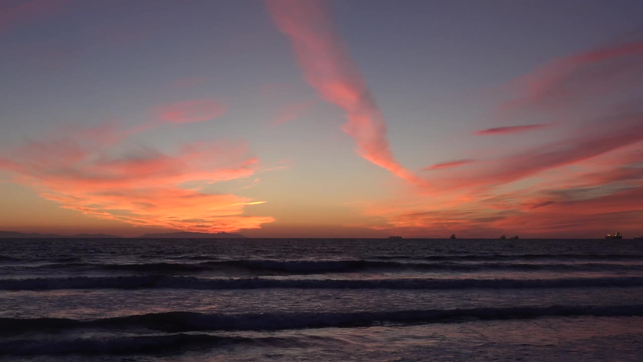 Huntington Beach, California shoreline at sunset, with a long pink cloud streak crossing the upper sky, broken wave lines in front, distant ships, and no visible people.