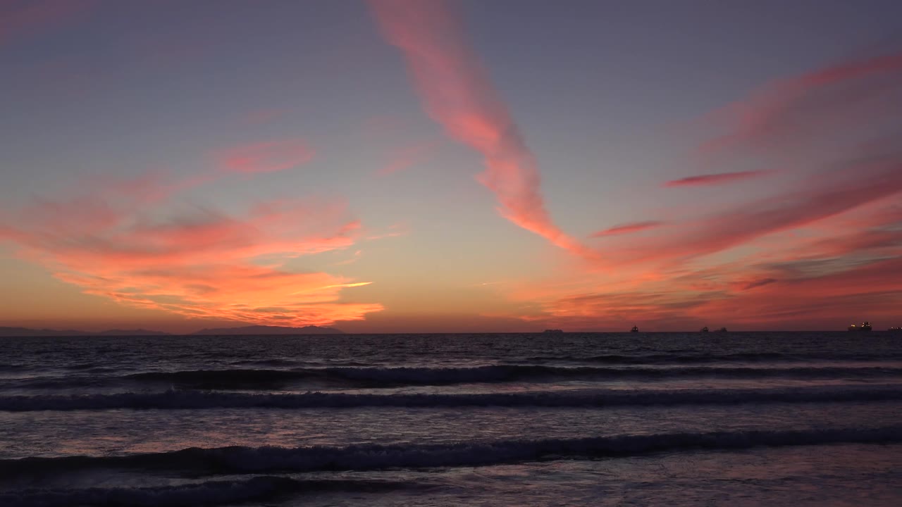 Huntington Beach, California dusk frame with the orange horizon low on the left, dark wave sets across the foreground, ship silhouettes offshore, and no visible people.