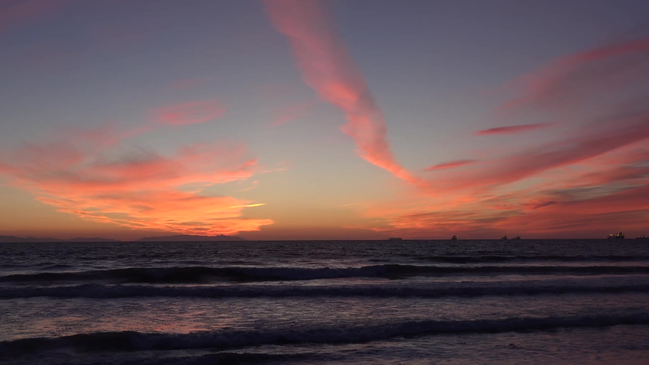 Huntington Beach, California sunset scene with rolling surf in shadow, a warm band of light near the horizon, pink cloud shapes overhead, ship silhouettes, and no visible people.