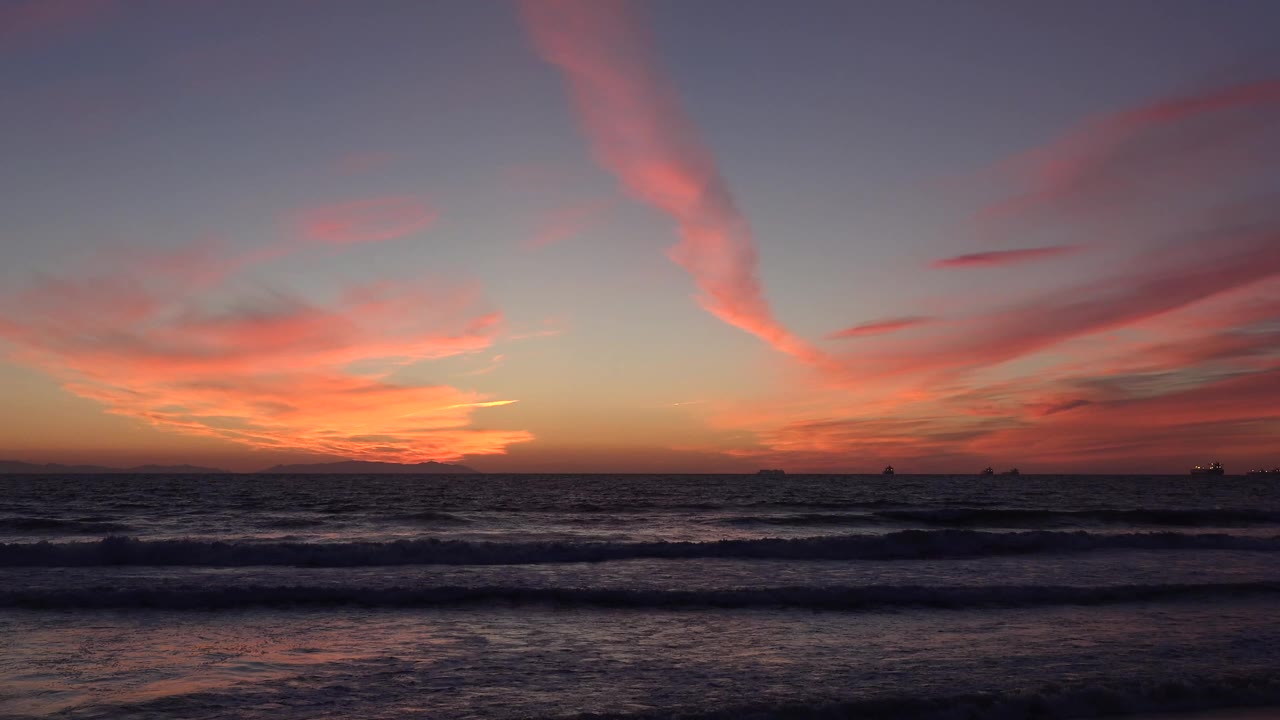 Huntington Beach shoreline at 0:01 with low waves advancing beneath a pink-orange sunset sky and no visible people.