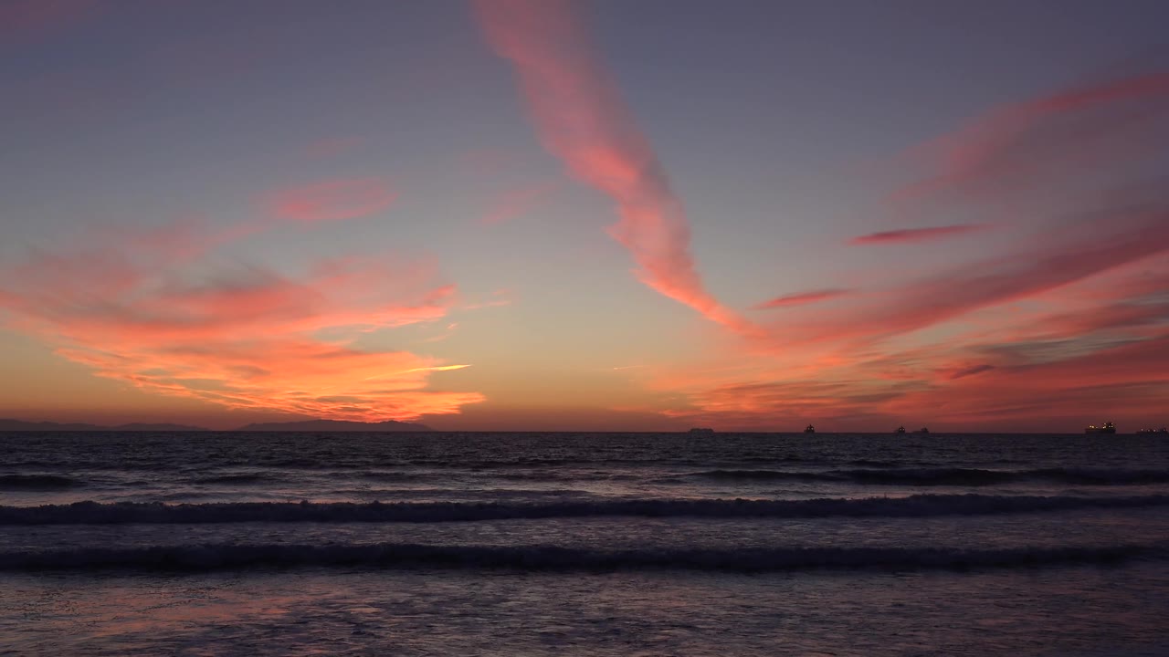 Huntington Beach at 0:03 with a wide pink cloud plume crossing the sky above steady shore waves and no visible people.