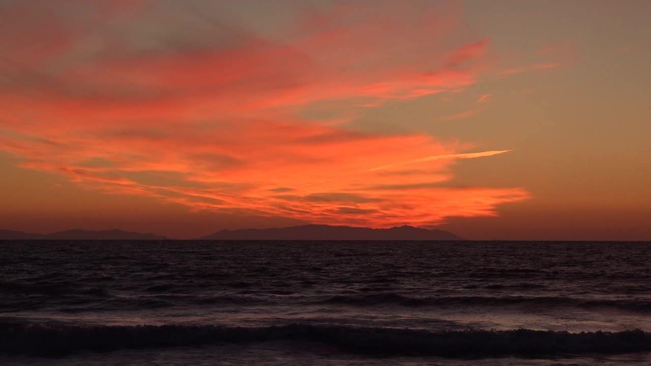 Orange sunset over dark ocean waves and distant shoreline silhouettes.