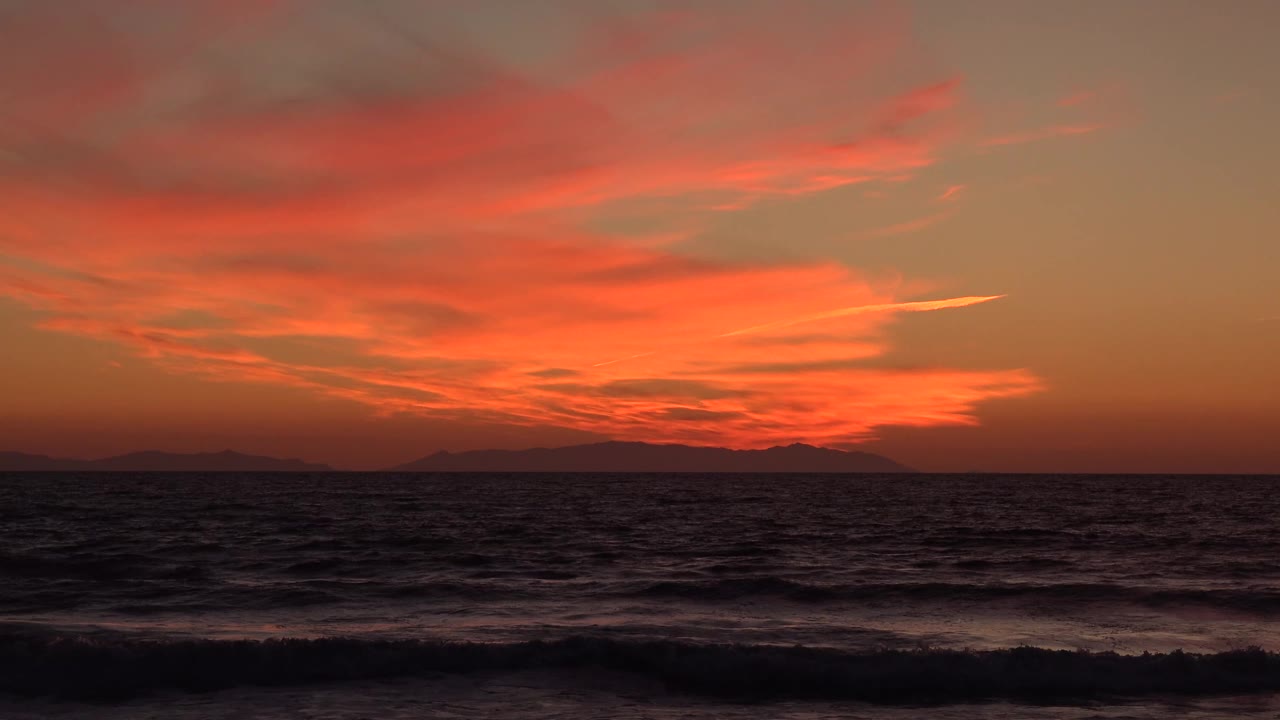 Huntington Beach ocean view at 0:01 with the surf line sitting low in frame, distant land in silhouette, brighter orange cloud detail near the center, and no visible people.