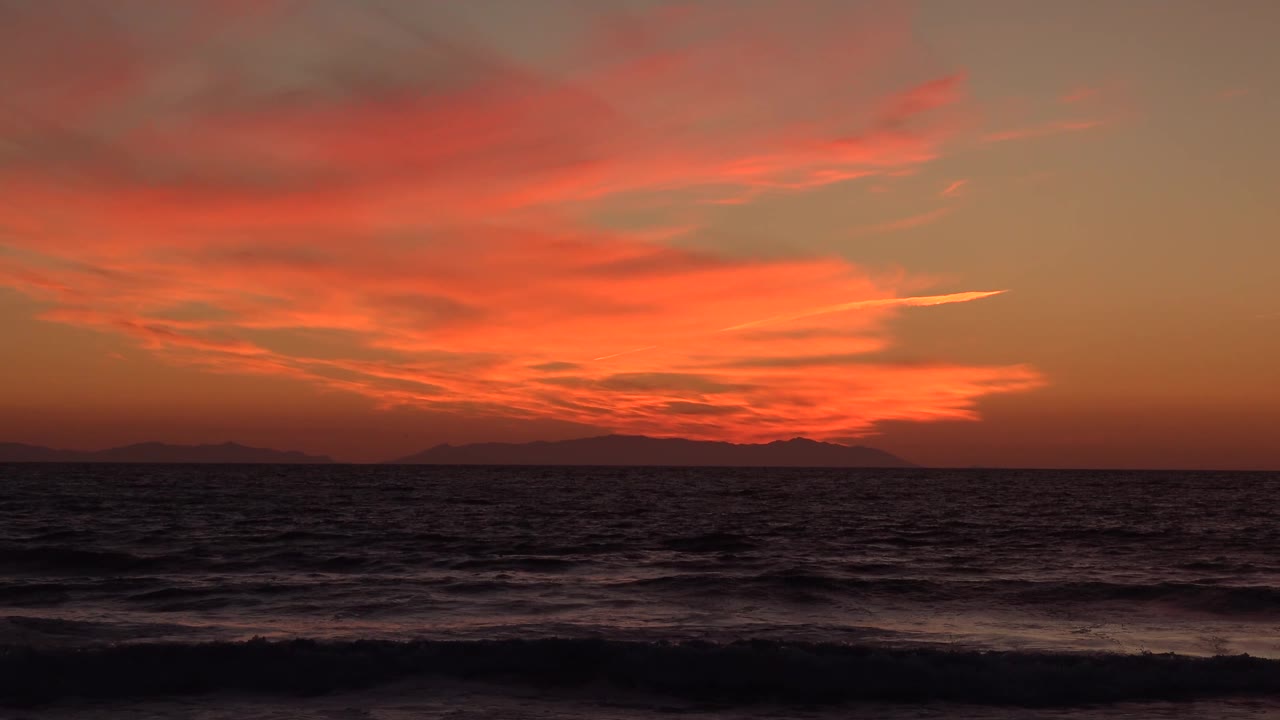 Huntington Beach sunset frame with a broad orange cloud bank, blackened water and small wave bands below, distant shore silhouettes, and no visible people.