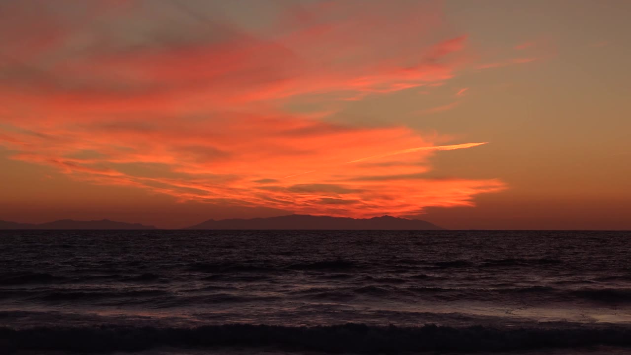 Huntington Beach shoreline view at 0:03 with choppy dark water in the foreground, a low band of silhouetted land, layered red-orange clouds, and no visible people.