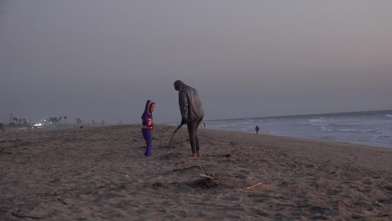 At Huntington Beach, two people face each other on dark sand near the surf under a dim pink-gray sky, with driftwood in the foreground and beach lights in the distance.