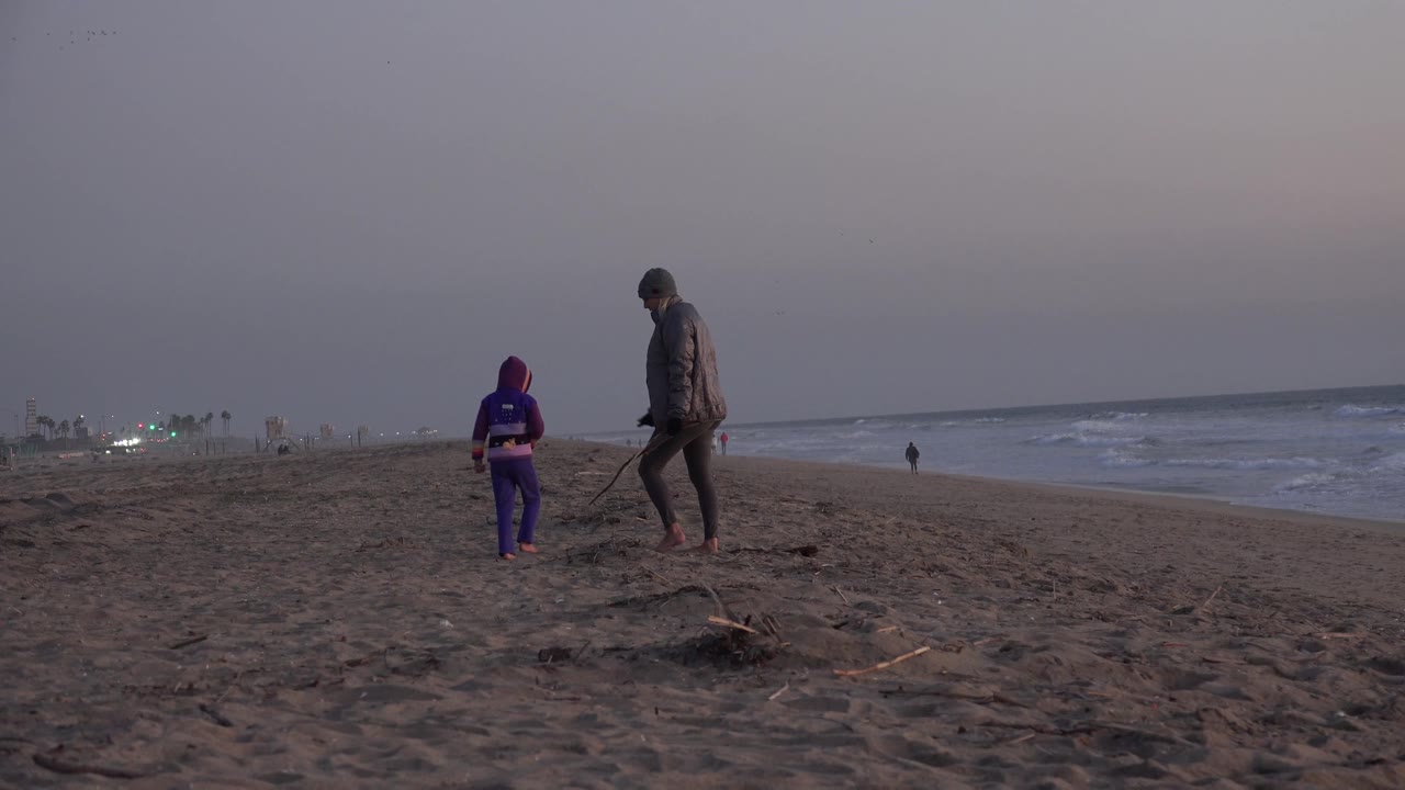 At Huntington Beach, the smaller person in purple and the taller barefoot adult walk away from the camera along dark sand, with surf on the right and faint lights on the left horizon.