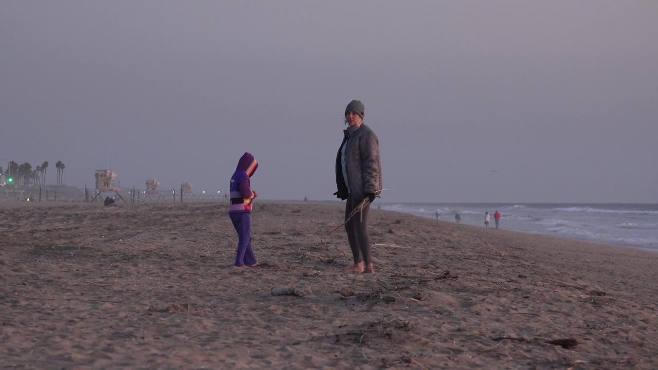 The child in purple stands left of the adult on the open Huntington Beach shoreline, while small waves break to the right and beach structures sit far in the haze.