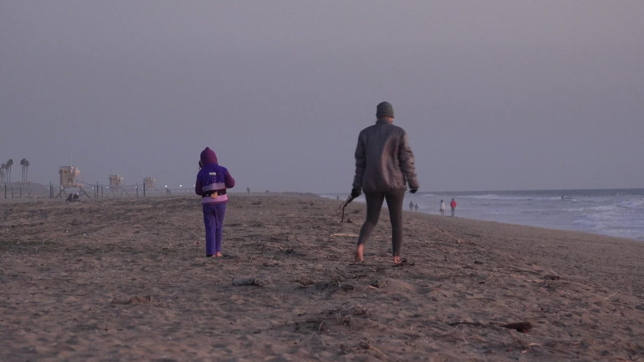 An adult and a child walk away from the camera on the sand at Huntington Beach, California, with waves on the right and distant beach structures on the left.