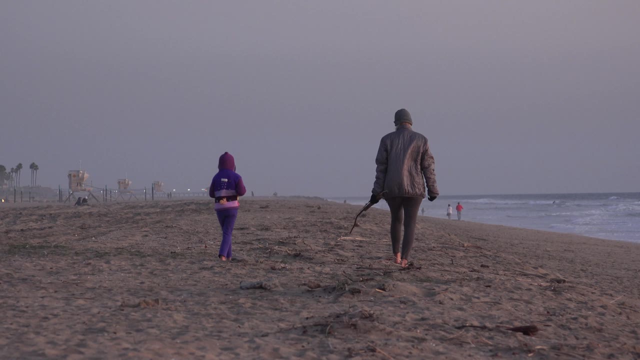 The adult and child are farther along the Huntington Beach sand, seen from behind with ocean waves at right and small figures near the water in the distance.