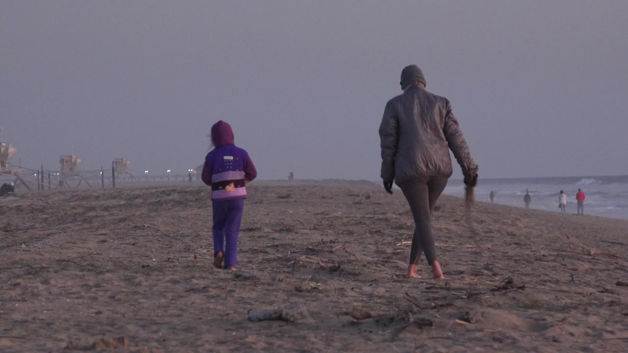 At Huntington Beach, California, a child in purple and an adult in a dark jacket walk away from the camera across dark sand with low surf and beach towers in the distance.