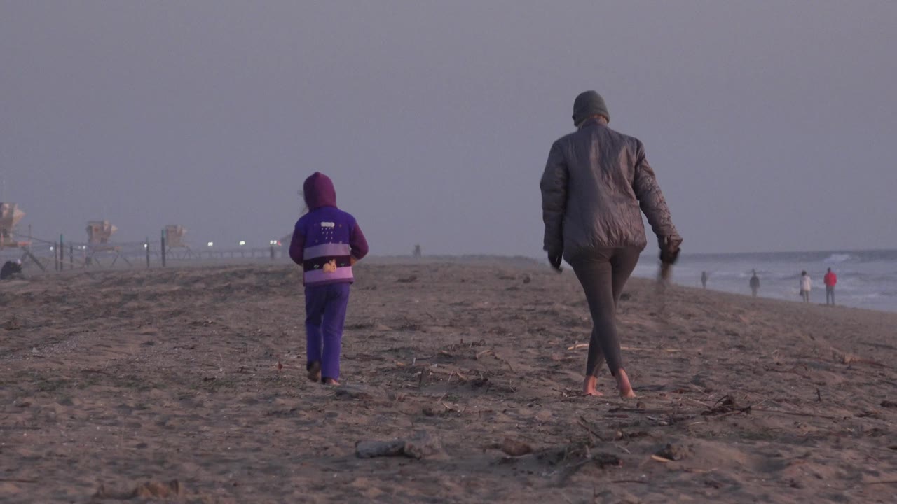 The child and adult move farther along the Huntington Beach sand, with the child left of center, the adult near center, and a pale surf line running along the right side of frame.