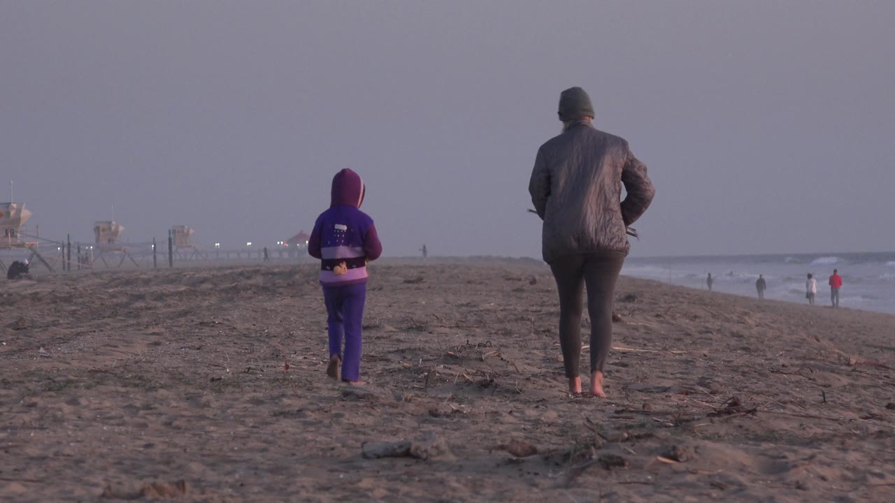 A rear view at Huntington Beach shows the adult slightly right of center and the child farther left, both walking barefoot over uneven sand while distant figures stand near the waterline.