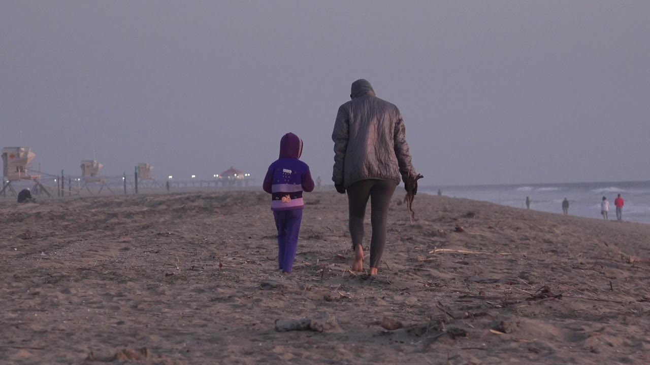 An adult in a hooded jacket and a child in purple walk away on the sand at Huntington Beach, with surf and distant beachgoers to the right.