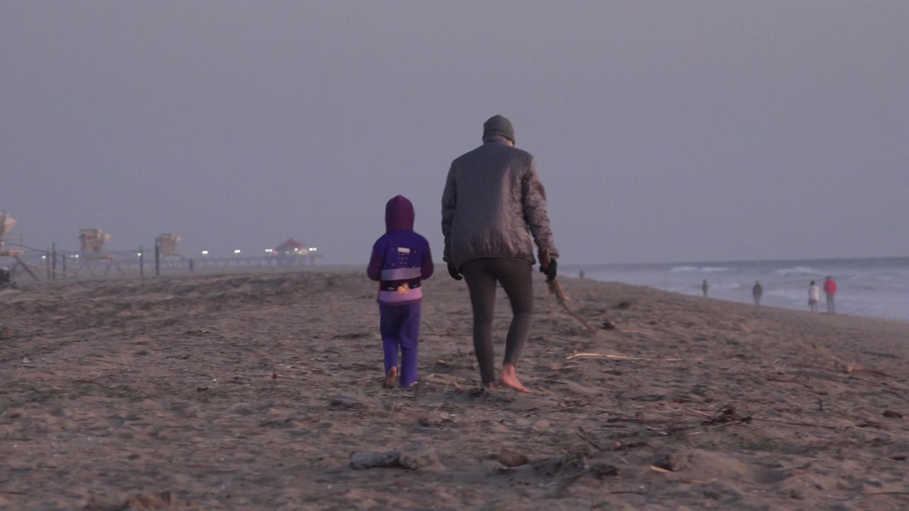 At Huntington Beach, the adult and child shift slightly right in the frame while waves break along the shoreline and a few distant people stand near the water.