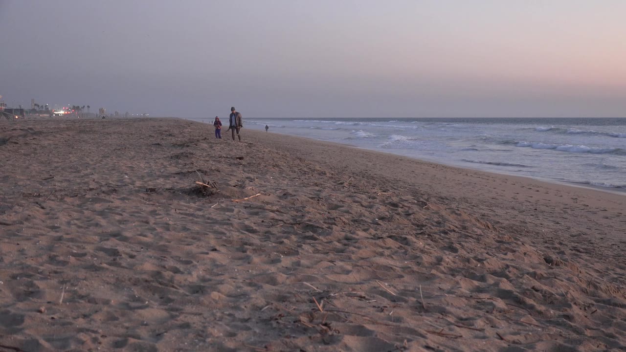 Huntington Beach shoreline with two distant walkers near center, smaller figures farther down the sand, and waves breaking along the right edge.