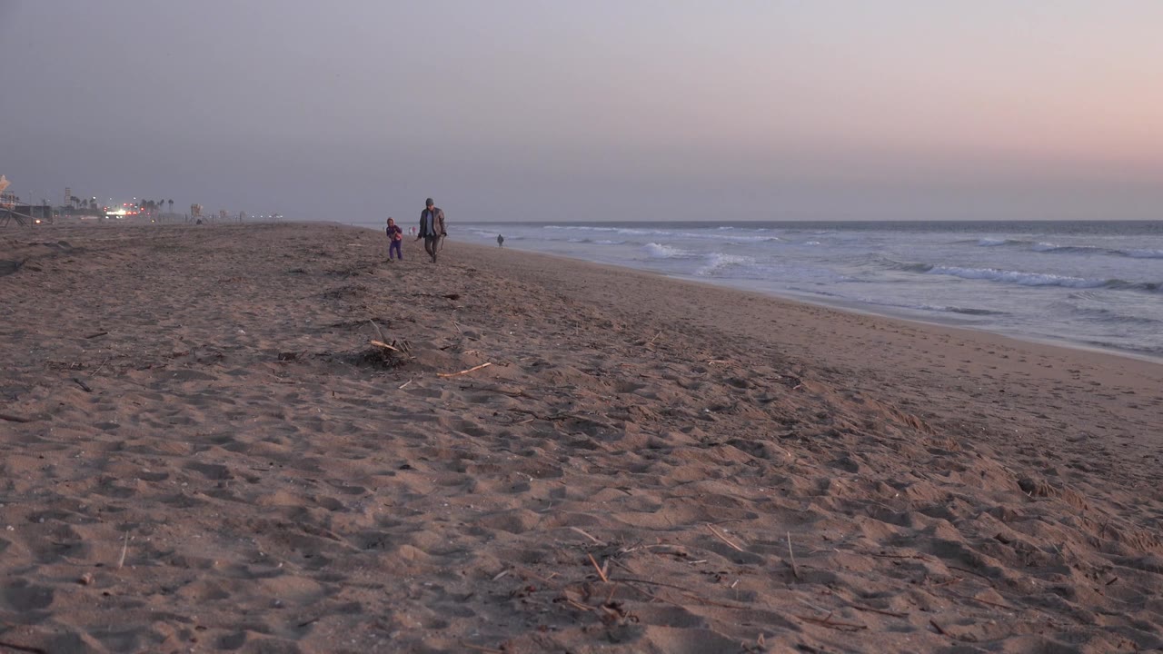 Huntington Beach at dusk with walkers spaced along the sand, a low wave line to the right, and soft pink light above the horizon.