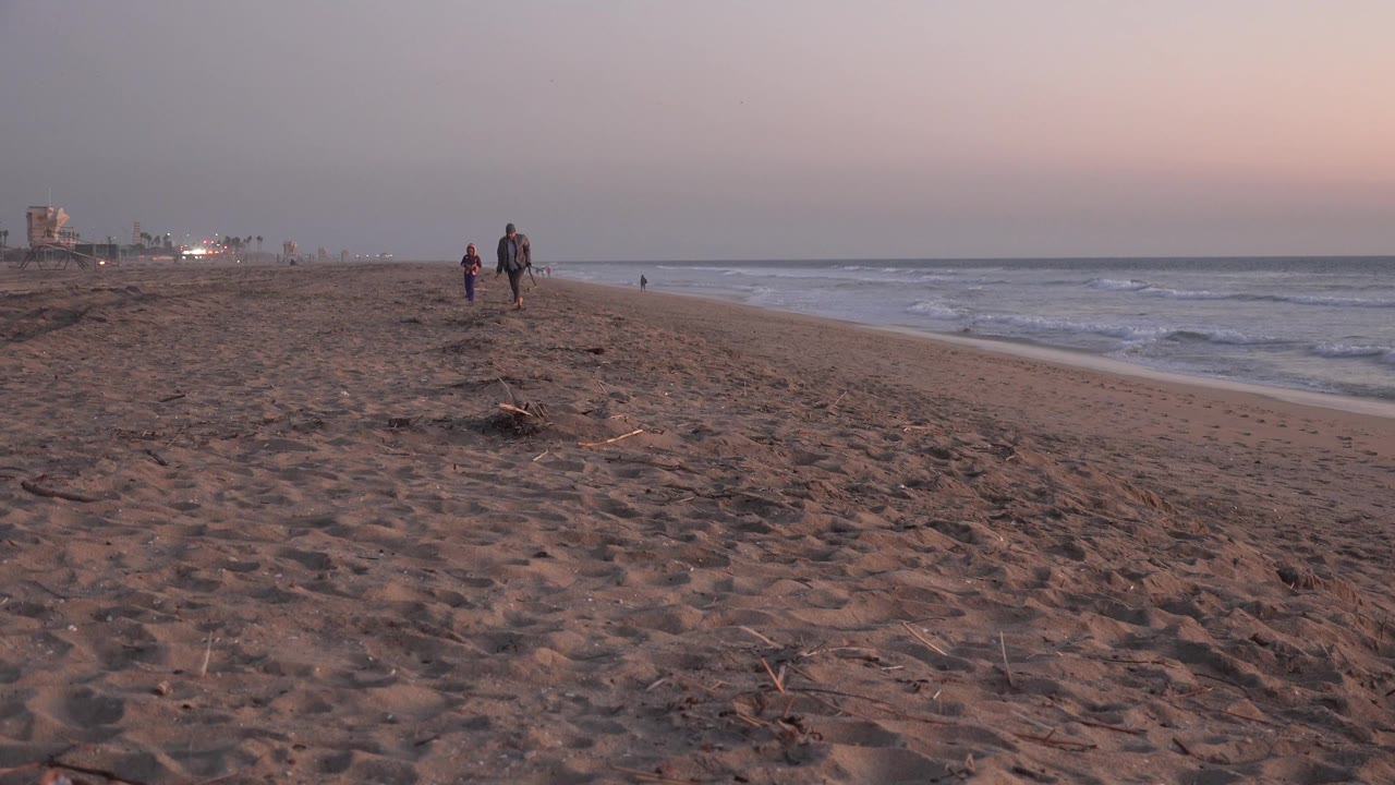 The two walkers remain in the midground on Huntington Beach, with rippled sand in front, low surf to the right, and muted sunset color above the horizon.
