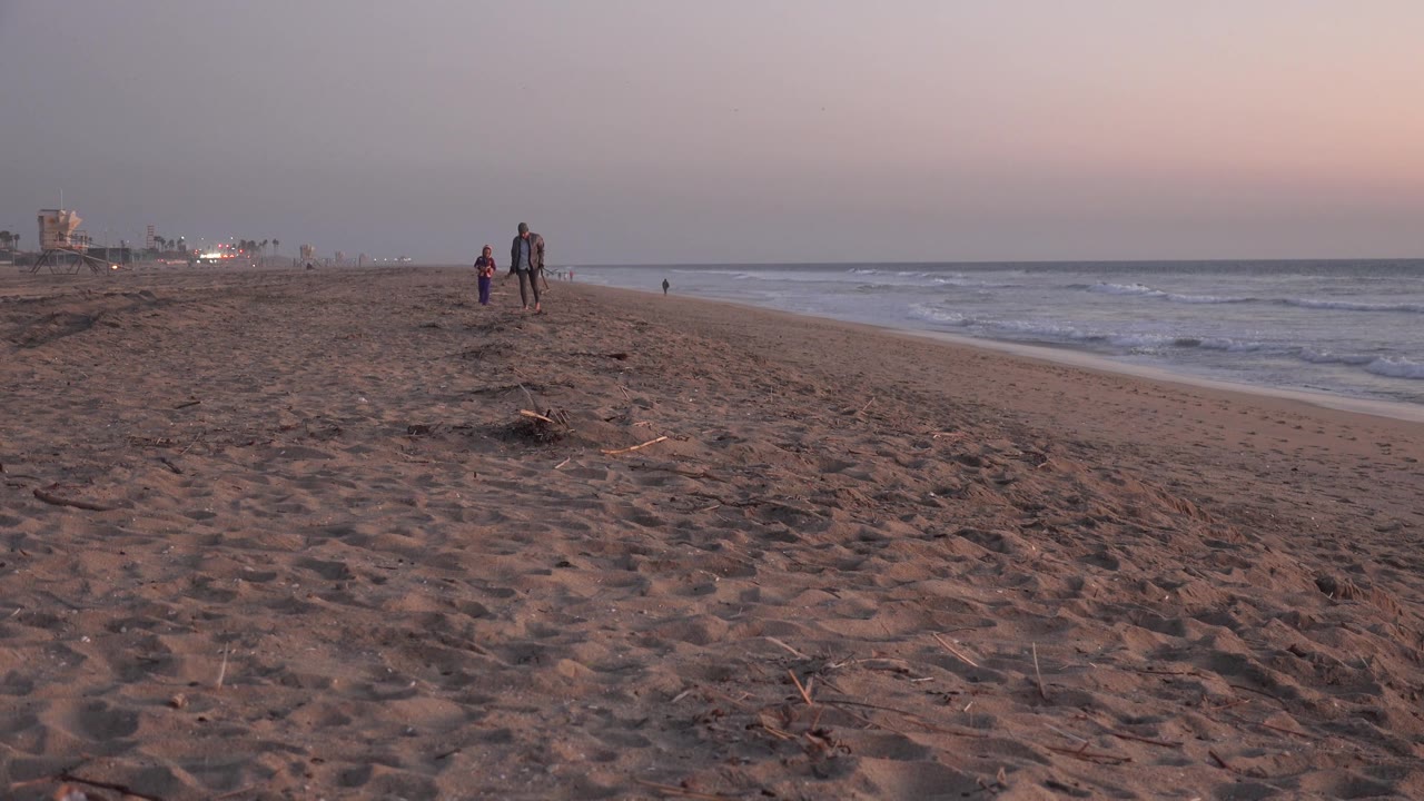 Two people continue approaching along the Huntington Beach shoreline, with the beach stretching diagonally to the surf and dim coastal structures far left.