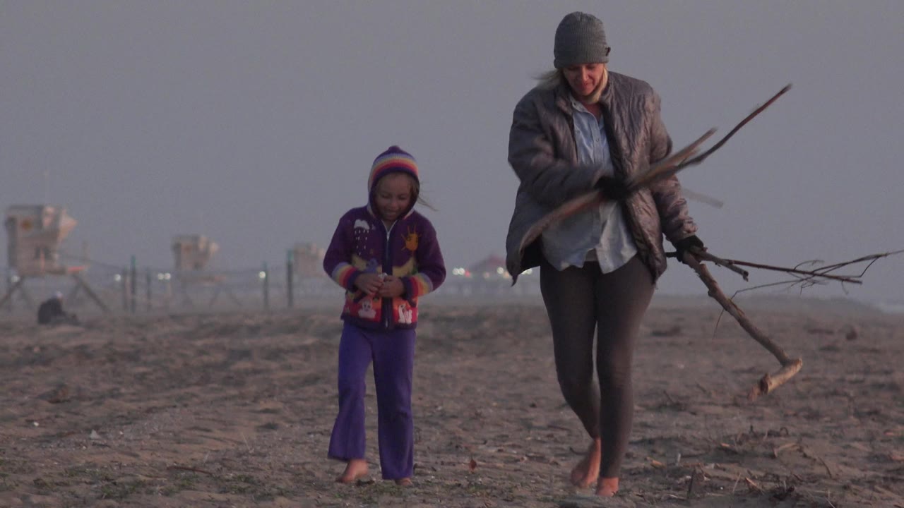 At Huntington Beach, California, a woman in a gray beanie carries branches while a child in purple walks beside her across the muted sandy shoreline.