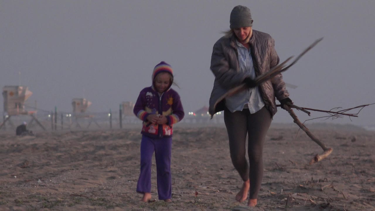 Woman holding branches walks beside child on beach.