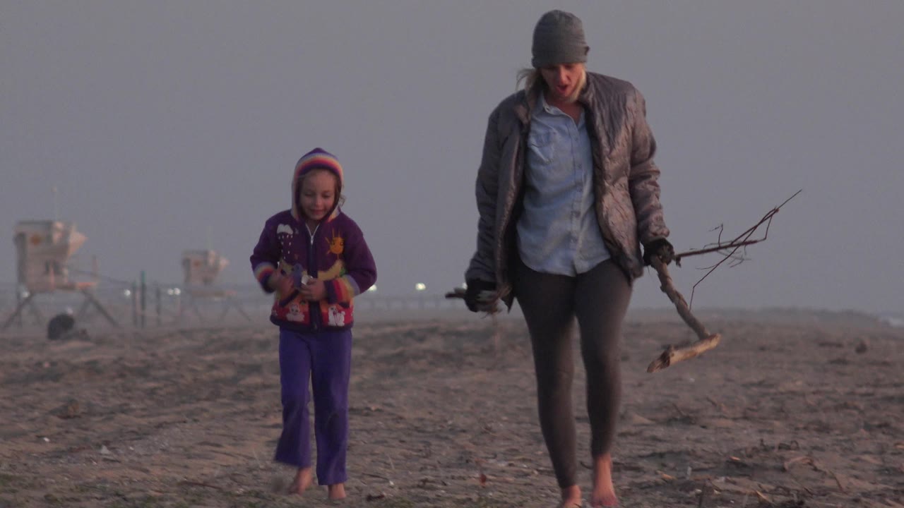 At Huntington Beach, California, the child in a purple outfit is farther left while the woman carrying branches moves closer to the right edge on the hazy shore.