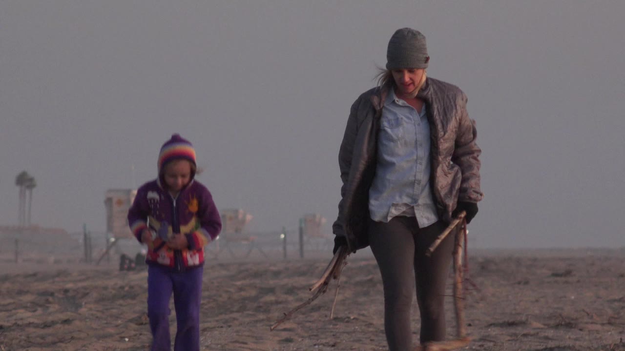 At Huntington Beach, California, a woman in a gray beanie and a young girl in purple walk on the sandy shore while carrying branches, with a hazy background behind them.