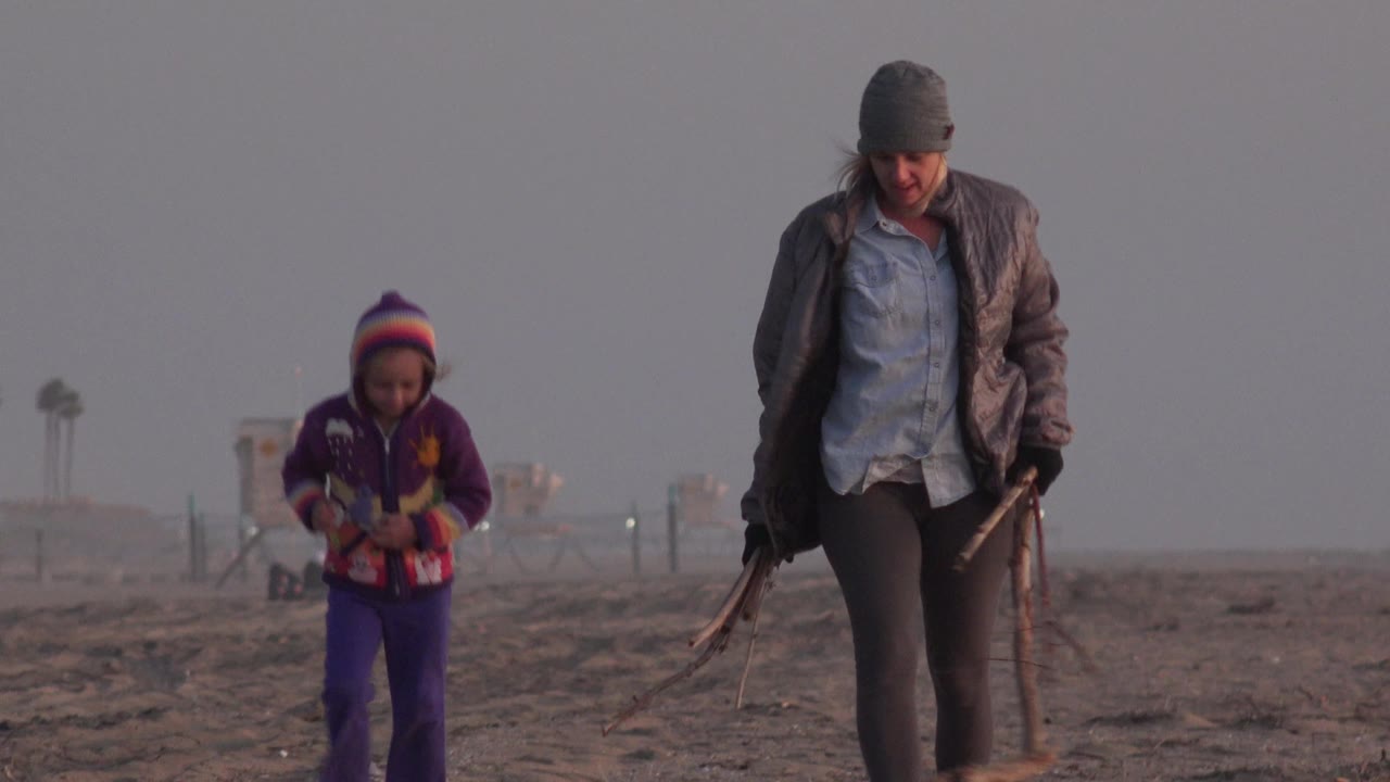 Young girl and woman walking separately across a gray beach scene.