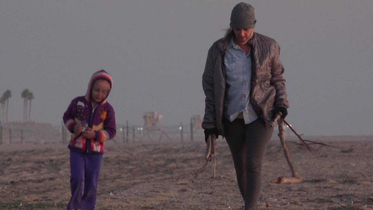 A child in a purple hooded outfit and a woman in a dark jacket continue across the Huntington Beach shore, with small traffic lights and faint structures visible in the haze.