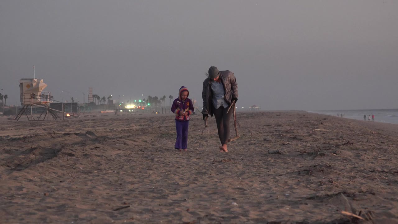 At Huntington Beach, California, a barefoot man carrying sticks walks beside a child on dark sand with a lifeguard tower and distant lights behind them.