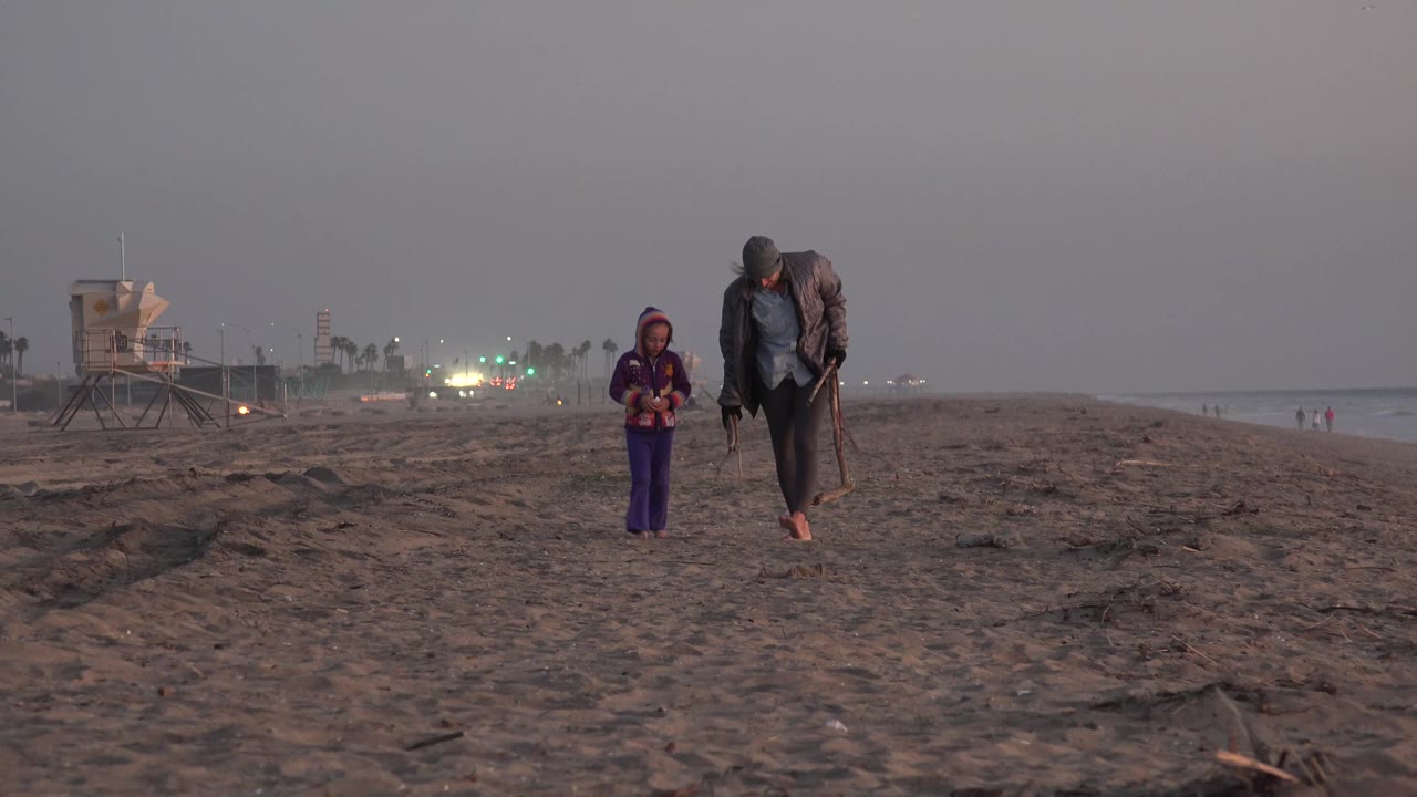 At Huntington Beach, California, the man and child continue across the open sand, with the ocean line to the right and beach lights glowing in the distance.